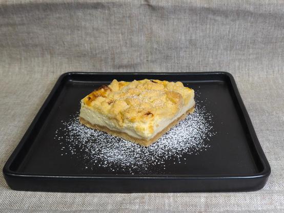 A piece of a special version of a German cheesecake - Badischer Käsekuchen - in the center of a black rectangular plate with icing sugar sprinkled around.