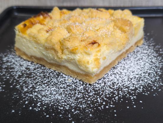 Close up of the piece of a German cheesecake with the focus on the front corner of the piece (the piece is in a 45 degree angle on the plate) in the center of a black rectangular plate with icing sugar sprinkled around.