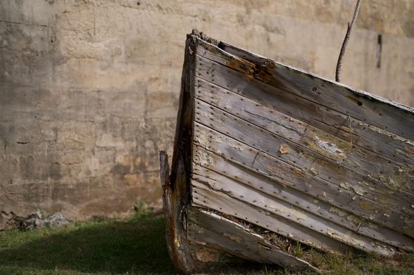 Photograph of an old wooden rowboat in front of a stone wall.  The boat has not been used for many years and the wood is partially rotted away.  Grass is growing through holes in the hull.