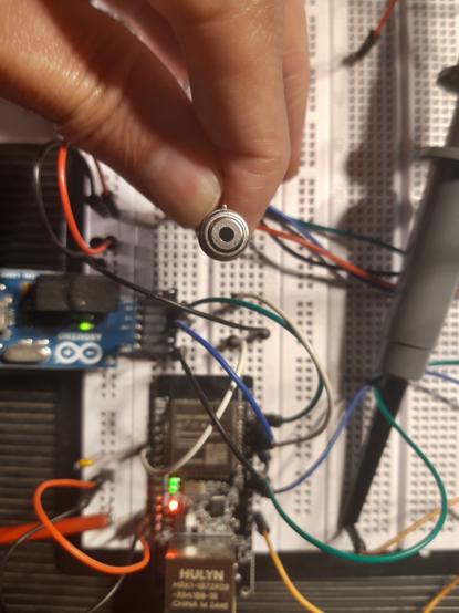 A top view of an electronics breadboard; a hand holds a circular sensor with four wires and a small black window in front of it, two small circuit board that are plugged into the breadboard are a bit out of focus.