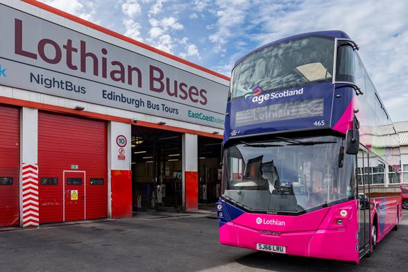 The image shows a bright, modern double-decker bus parked outside the Lothian Buses depot. The bus features a vibrant purple and pink colour scheme with the “Age Scotland” logo visible on the front above the windscreen. It is branded “Lothian” with fleet number 465 and bears the registration plate SJ66 LRU. The depot building, mainly red and white, has large roller shutters, one of which is open. Above the doors, a large sign reads “Lothian Buses”, with additional services listed, including NightBus, Edinburgh Bus Tours, and EastCoastbuses. A small road sign indicates a speed limit of 10 mph.
Photo by Lothian Buses ltd.