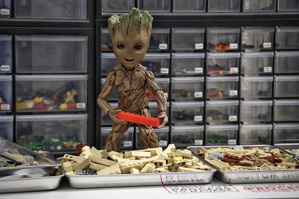A toy Groot holds a red Lego brick, seemingly about to set it on a pile of tan Lego bricks in from of him. There are many more Lego bricks all around.