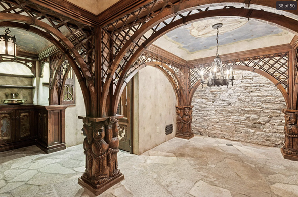 A basement with medieval style wooden arches with what looks like monk figures carved into the bases. There is stonework on the walls and floor and a chandelier hanging in the middle.