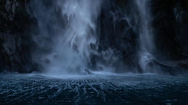 Detail photo of the bottom of a waterfall. The area around it is dark and the water beneath it has small waves of water that spread out like a blue grid on black. The tones are white, blue, and black.