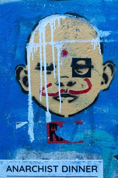 color photograph, portrait format. a house wall, a stencil. a beige-yellow face with black contours in front of a blue background. white lines over parts of the face. sticker, red. at the bottom a poster cutout with the words: “anarchist dinner”.

farbfotografie, hochformat. eine hauswand, ein stencil. ein beige-gelbes gesicht mit schwarzen konturen vor blauem hintergrund. weiße linien über teilen des gesicht. aufkleber, rote farbe. am unteren rand ein plakatausschnitt mit den worten: "anarchist dinner".