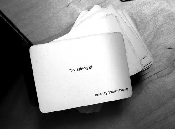 A black-and-white photo of an Oblique Strategy card. The card reads: 'Try faking it! (given by Stewart Brand)'.