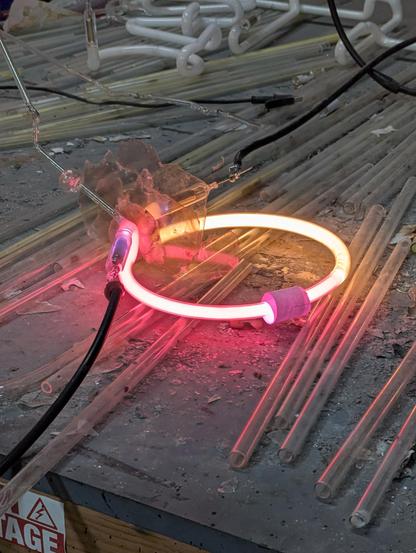 My assistant bombarded this tube! So I could get this shot- you can see the setup- mercury trap bubble in the clear tube in the side of the pink double back bend. There is a bunch of mica to prevent short circuiting and destroying the rube and high power bombarder leads are hooked up to the electrodes- for overpowering and purifying the tube. Pink and warm white tube on a table full of glass and ash.