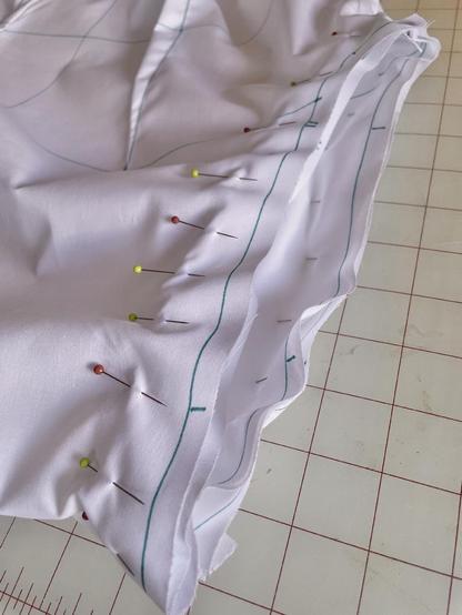 Closeup of a set-in sleeve pinned into the armscye of a shirt made of plain white cotton. Its seam lines and alignment notches are marked in teal pen.