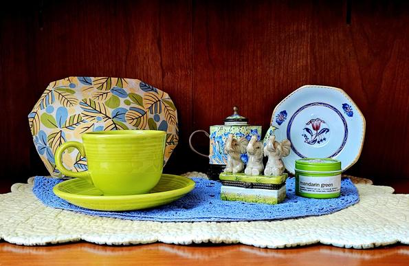 Tea tableau in yellow, blue, and bright green, centering around a small ceramic figural trinket box in the shape of three seated grey elephants raising their heads. There is a vintage Fiestaware cup and saucer, and enameled copper miniature Kelvin Chen teapot, and antique saucer, and floral tea cozy, all on a blue crochet doily atop a yellow quilted runner.