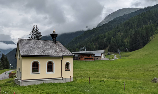 A small chapel with beige walls and black shingle roof, in a green mountain valley with a ski lift in the background. According to legend, a supernatural horse appeared here at night which would quickly transport herders to their lovers and back again - at the cost of their souls.