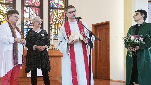 Ein Mann mit hellem Talar und roter Stola spricht stehend in ein Mikrofon. Zwei Frauen stehen links hinter ihm, eine weitere mit Kollarhemd rechts neben ihm. Im Hintergrund ein Abendmahlstisch und drei Buntglasfenster.