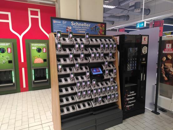 Handscanner in der Ausgabestation, daneben ein Kaffeeautomat, dahinter Pfandautomaten.