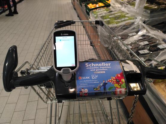 Handscanner am Einkaufswagen. Letzterer ist beschriftet mit „Schneller einkaufen, bezahlen und genießen. Einfach mehr drin! K-Scan. 1. Werde Kaufland Card Mitglied. 2. Scanne deine Produkte während des Einkaufs. 3. Bezahle an der SB-Kasse.“