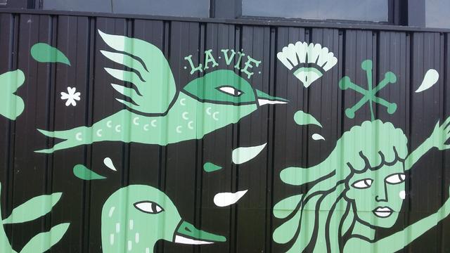 Motif en vert sur fond noir : oiseau, tête féminine, tête de canard. Texte : la vie