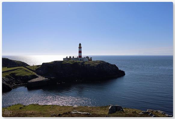 Ein rot-weißer Leuchtturm steht auf einer Landspitze. Der Himmel ist blau, wolkenlos. Die Sonne scheint, das Meer glitzert. Im Vordergrund grünes Land mit Felsen.

A red and white lighthouse stands on a headland. The sky is blue, cloudless. The sun is shining, the sea glistens. Green land with rocks in the foreground.