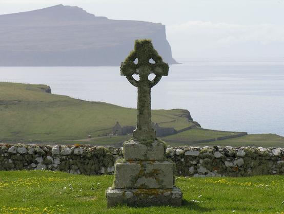 The graveyard at Trumpan Church. The image shows a mossy gravestone in the centre foreground comprising a Celtic cross on a plinth. It is set in grass and there is a stone wall behind it. Beyond that the ground drops away steeply giving a view of two buildings on a shoreline with sea beyond, and a promontory coming in from the left with cliffs in the distance. The scene is in sunlight.