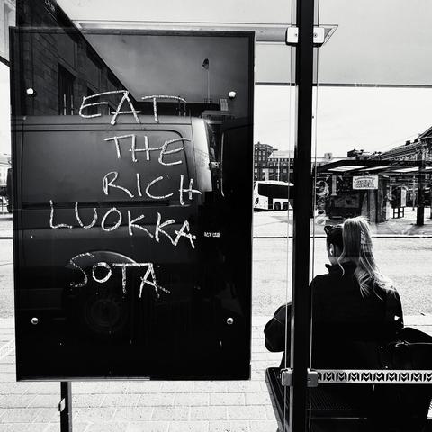 A bus stop display board shows graffiti reading "EAT THE RICH LUOKKASOTA," while a woman sits casually in silhouette nearby.