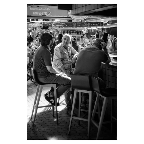 This is a portrait format black and white image of three men sitting at a bar in the central market in Valencia. Sitting on high stools, the two men in the foreground are in shadow with the one to the right with his back to the viewer and the one to the left side on. The man in the centre faces the viewer and is lit from above by sunlight falling through the market's glass roof. Taken with a Sony RX100iii.