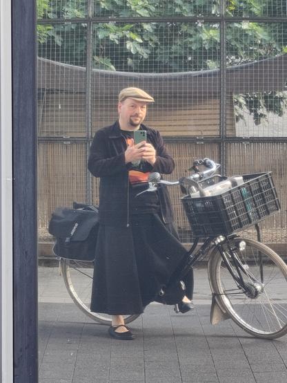 Selbstporträt in spiegelnder Scheibe. Ich sitze auf meinem Fahrrad mit einem Fuß auf dem Boden. Ich trage eine schwarze Jacke, einen langen weiten schwarzen Rock, schwarze Ballerinas mit Schnürung, ein schwarzes T-Shirt mit orangefarbenem Aufdruck (Fury in the Slaughterhouse - Hope Tour 2023), und eine mittelbraune Schirmmütze. Das Fahrrad ist ein schwarzes Hollandrad mit Schwanenhals-Rahmen, einem großen Korb am Lenker und Packtaschen am Gepäckträger.