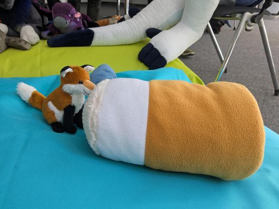 Photo of a small red fox plush, sitting in front of a plush beer mug that's lying sideways, and pushing its nose against it. Both plushies are on a light blue pillow, with other plushies and parts of a panel room visible in the background.