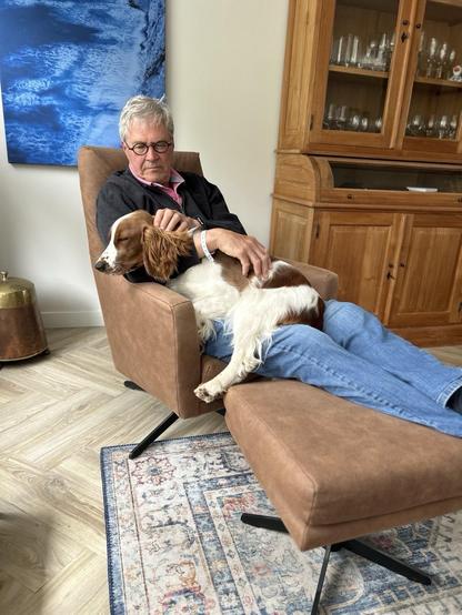 An older man with glasses sits in a brown chair, gently holding a resting dog on his lap. The background features a blue artwork, wooden furniture, and a patterned rug on the floor.