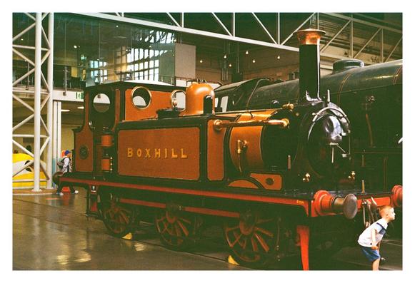 Inside a spacious railway museum, a beautifully preserved vintage steam engine named Boxhill gleams under warm lighting. The engine is painted in rich brown and black with brass and red details, its polished surfaces evoking the craftsmanship of a bygone era. To the right, a curious young boy in a white and blue shirt bends forward excitedly, peering closely at the front of the train with wide-eyed wonder. Behind the engine, modern glass walls and steel beams contrast with the locomotive's historic charm, bridging past and present in a vibrant celebration of engineering and childhood fascination.