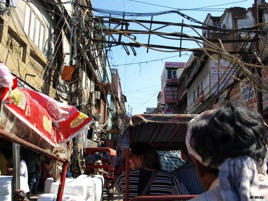 Farbfoto, aufgenommen in einer der engen Straße von Old Delhi, Indien. Rechts vorn der Kopf eines Rikschafahrers von hinten, davor eine Rikscha mit Leuten. Links ein Laden mit weißer Ware und rotem Banner. Häuser links und rechts, verbunden durch ein Metallkonstruktion mit vielen Kabeln.