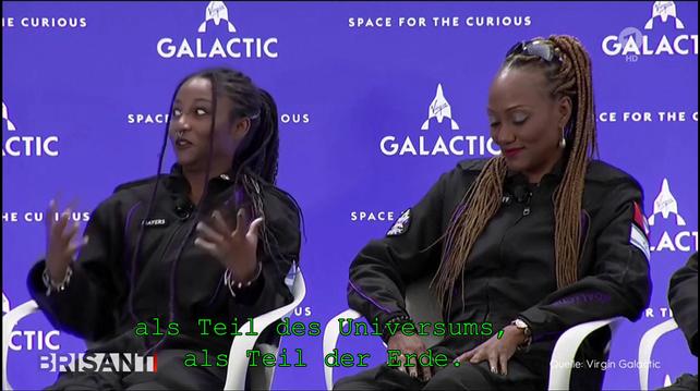 Description: The image shows two women sitting on chairs in front of a blue wall, both wearing black dresses. On the wall behind them is a banner with the words "Space for the Curious" written on it.

Disclaimer: This image description was automatically generated and may not be entirely accurate.