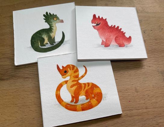 three mini dragon paintings. One is a dark and light green one with yellow eyes, one is a chubby red and pink striped one and the other is dark and light orange with beady black eyes and a long tail.