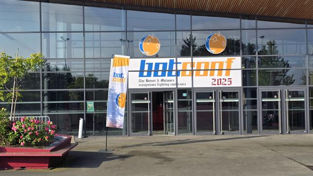 View of a conference hall entrance, with glass windows surrounding the door. A Botconf 2025 banner is printed above the doors and a vertical banner is standing in front to the left. A bench with flowers can be seen to the far left of the image.