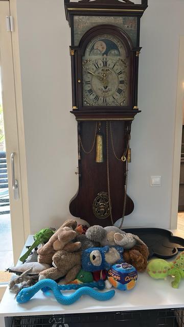 A vintage Dutch wall clock is shown on the left, while a pile of various stuffed toys, including a blue bear, several soft animals, and a colorful toy ball, is arranged on a white dog bench beneath it.