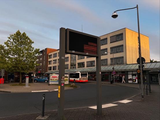Bahnhofsvorplatz mit Bushaltestellen, davor eine kaputte digitale Anzeigetafel, dahinter angrenzende Gebäude. Ein Bus ist im Hintergrund zu sehen, und es gibt Bäume und Menschen in der Nähe. Am Himmel ein paar Wölkchen und Abendlicht.