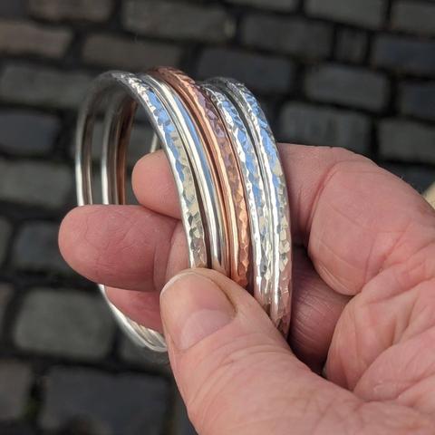 Five chunky bangles (four silver, one copper in the middle) being held between the fingers of a hand. Most of the bangles have a hammered texture. In the background are street cobbles.