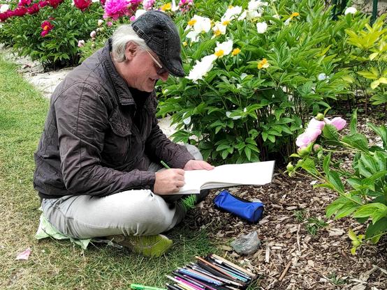Reinhard sitzt am Boden vor einer Pfingstrose mit grossen rosa Blüten. Er zeichnet eine Blüte mit Farbstiften die man vor ihm am Boden ausgebreitet sieht.