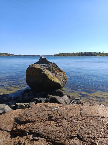 Picture of a large rock on the edge of the sea, with a view to the archipelago behind it, and clear blue skies.