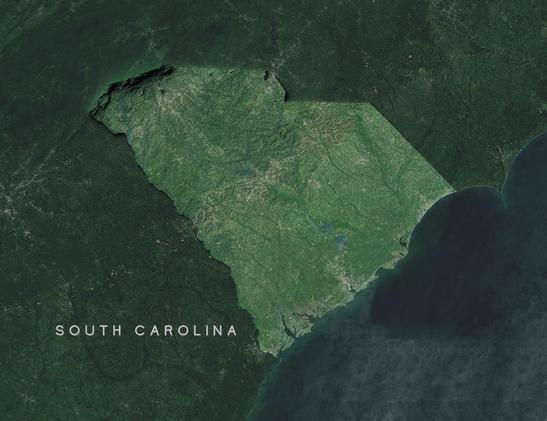 Satellite topographical map of South Carolina. The state outline is highlighted against a dark green background. The image shows the state's varied topography, with the Blue Ridge Mountains visible as darker terrain in the northwest corner, transitioning to the rolling Piedmont region in the center, and the flat coastal plain in the southeast. The Atlantic Ocean appears as dark blue along the eastern border. Several river systems are visible cutting through the landscape. Urban areas appear as lighter patches, with the Charleston area visible along the coast. The map effectively illustrates the state's geographic transition from mountains to sea.