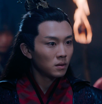 Close up of Wen Chao in the Xuanwu's cave, an expression of anger on his face. A flaming torch is visible being held by someone behind him off toward the right side of the image.