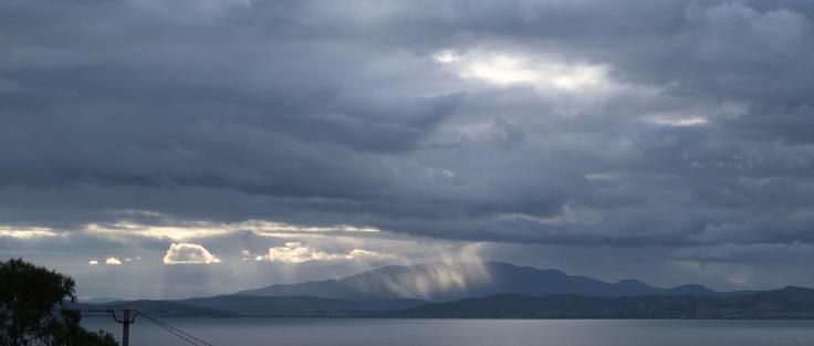 A moody sky fills most of the frame in the late afternoon.  The sun beams down through a distant cloud gap, as rain falls in front of a small mountain, the rain lit in the beam.  Hills lie in semi silhouette along the horizon, a large bay in the foreground reflecting the steel blue sky.