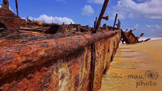 This image shows the rusted remains of a shipwreck on a sandy beach. The metal structure is corroded and weathered, with visible bolts and rust patterns. The ocean and sky are visible in the background, creating a coastal scene.