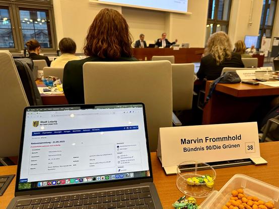 Foto aus der Ratsversammlung. Im Vordergrund mein Laptop, mein Namensschild sowie Schokolade und Erdnüsse. Im Hintergrund sind Stadtratsmitglieder sowie der OBM Burkhard Jung zu sehen.