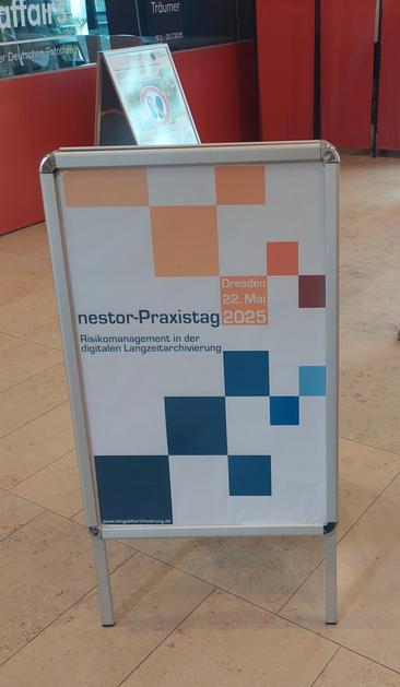 Ein aufgestelltes Plakat in einem Innenraum kündigt den "nestor-Praxistag" am 22. Mai 2025 in Dresden an. Das Thema der Veranstaltung lautet "Risikomanagement in der digitalen Langzeitarchivierung". Das Design des Plakats zeigt ein Muster aus farbigen Quadraten in Orange- und Blautönen. Unten ist die Website "www.langzeitarchivierung.de" angegeben.