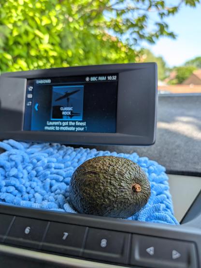 A ripe avocado rests on a blue microfiber cloth placed on a car dashboard. In the background, a digital car display screen shows a BBC Radio 6 Music station playing classic rock at 10:32 AM. Green foliage and houses are visible outside the car through the windshield.