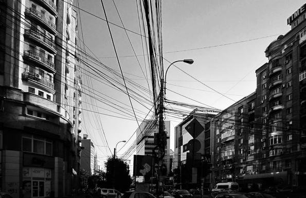 A black and white photograph captures a street scene dominated by a dense network of overhead wires. The wires crisscross the sky, creating a chaotic web against the backdrop of tall buildings on either side of the street. The buildings are multi-storey, suggesting an urban environment. A street lamp stands prominently amidst the wires. The lower part of the image shows cars and a van on the street, and there are some signs visible on the buildings, including one that reads "VICHY". The overall impression is one of a bustling city street, with the wires adding a distinctive, if somewhat cluttered, visual element.