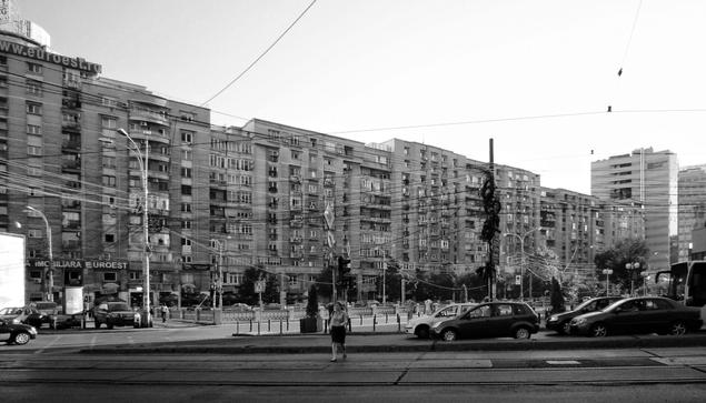 A black and white photograph captures a street scene in Romania, specifically Bulevardul 1 Decembrie 1918, characterised by communist-era architecture. The image shows a long row of multi-storey buildings, typical of the communist era, lining the street. Overhead wires crisscross the scene, adding a layer of visual complexity. Cars and a van are visible on the street. A sign reading "EUROEST" is visible on one of the buildings. The overall impression is of a densely populated urban area, with the architecture reflecting a specific historical period.