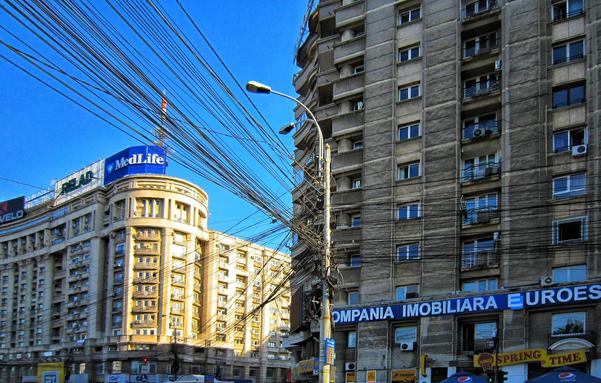 The image captures a street scene in Bucharest, Romania, near Piața Obor. A complex network of overhead wires dominates the view, crisscrossing against a clear blue sky. On the left, a building with a 'MedLife' sign is visible. To the right, a tall, communist-era building displays a sign reading 'COMPANIA IMOBILIARA EUROEST'. Street-level shops are visible with signs like 'SPRING TIME' and 'VELO ALMAJA'. The image offers a vibrant view of the city, with a mix of architectural styles and urban elements.