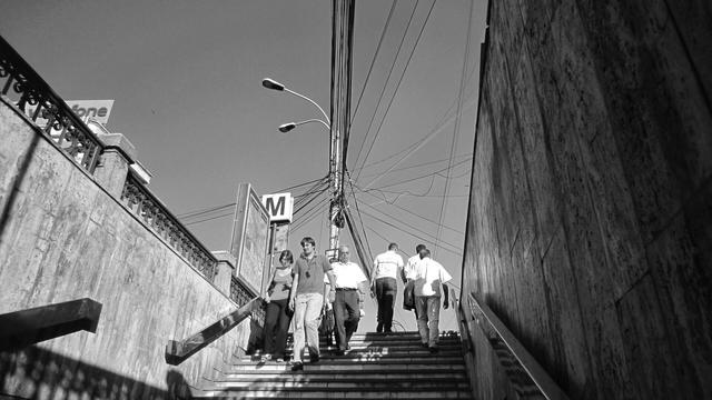 A black and white photograph shows a group of people walking down a set of steps, possibly an entrance to a metro station, indicated by a sign with the letter "M". Overhead wires are visible, crisscrossing against a clear sky. The scene is framed by concrete walls and a decorative railing. The image captures a moment of everyday urban life.