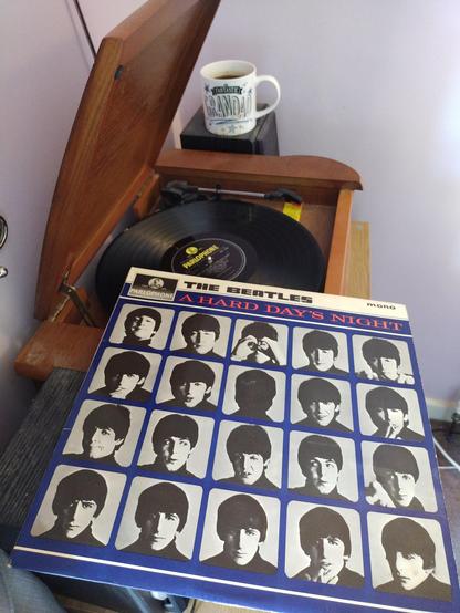 Record player with a 12" black vinyl record.
In the foreground is an album cover with four rows of five photographs each of John, George, Paul and Ringo.
Behind the player is a mug of coffee on a speaker.