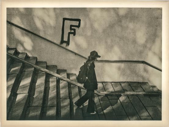 This image is a lith print photograph, characterized by its distinctive gritty texture, high contrast, and warm, often sepia-toned hues.

The scene captures a solitary figure—likely a young woman—ascending a wide, outdoor staircase. She is dressed in dark clothing, wearing a coat, a baseball cap, and a backpack. The staircase and the adjacent wall create strong diagonal lines that guide the viewer's eye through the composition. On the wall, the letter "F" is boldly spray-painted in a stylized, graffiti-like font.

Soft, dappled light filters across the scene, likely from trees casting shadows, adding a dreamlike or abstract quality. The use of lith printing enhances the mood: the shadows are deep and textured, while the highlights retain a delicate softness. The borders of the image are slightly darker and warm-toned, typical of prints on aged or fiber-based paper.