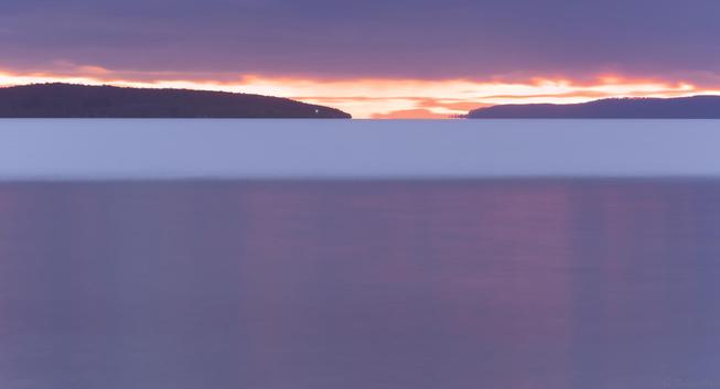 A long exposure sunset photo.  The lower half is darker glassy water in the foreground, with pale pink streaks where wavelets catch the light.  Intermediate to distant waters are wind affected, changing the surface colour to a pale grey blue.  Land lays in silhouette across the water.  A brilliant orange sunght glows in a thin line on the horizon, low darker cloud cover above.