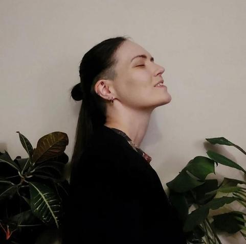 Profile picture. A youngish woman with black clothes and black hair, looking happily sideways. Surrounded by plants.